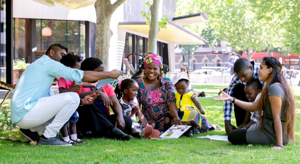 A group of smiling multicultural people sit on the grass outside Bendigo Library