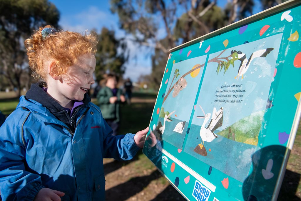 Young girl with red, curly hair smiles and points at a StoryWalk sign