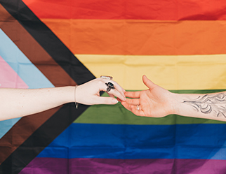 Two hands reaching to touch eachother in front of a progress pride flag