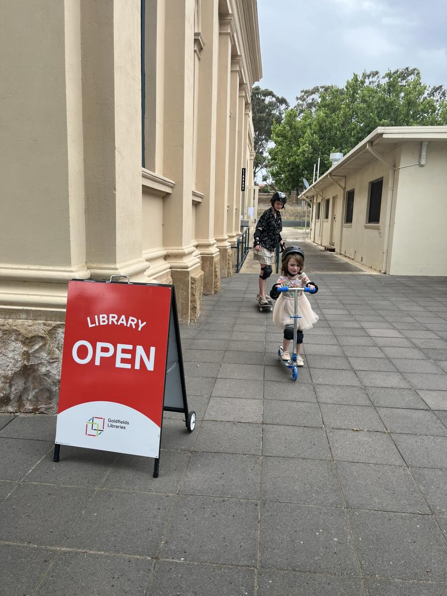 two children using sports equipment from Heathcote Library. In the background, a teenage boy rides a skateboard. In the foreground, a younger girl rides a scooter. Both are wearing helmets.