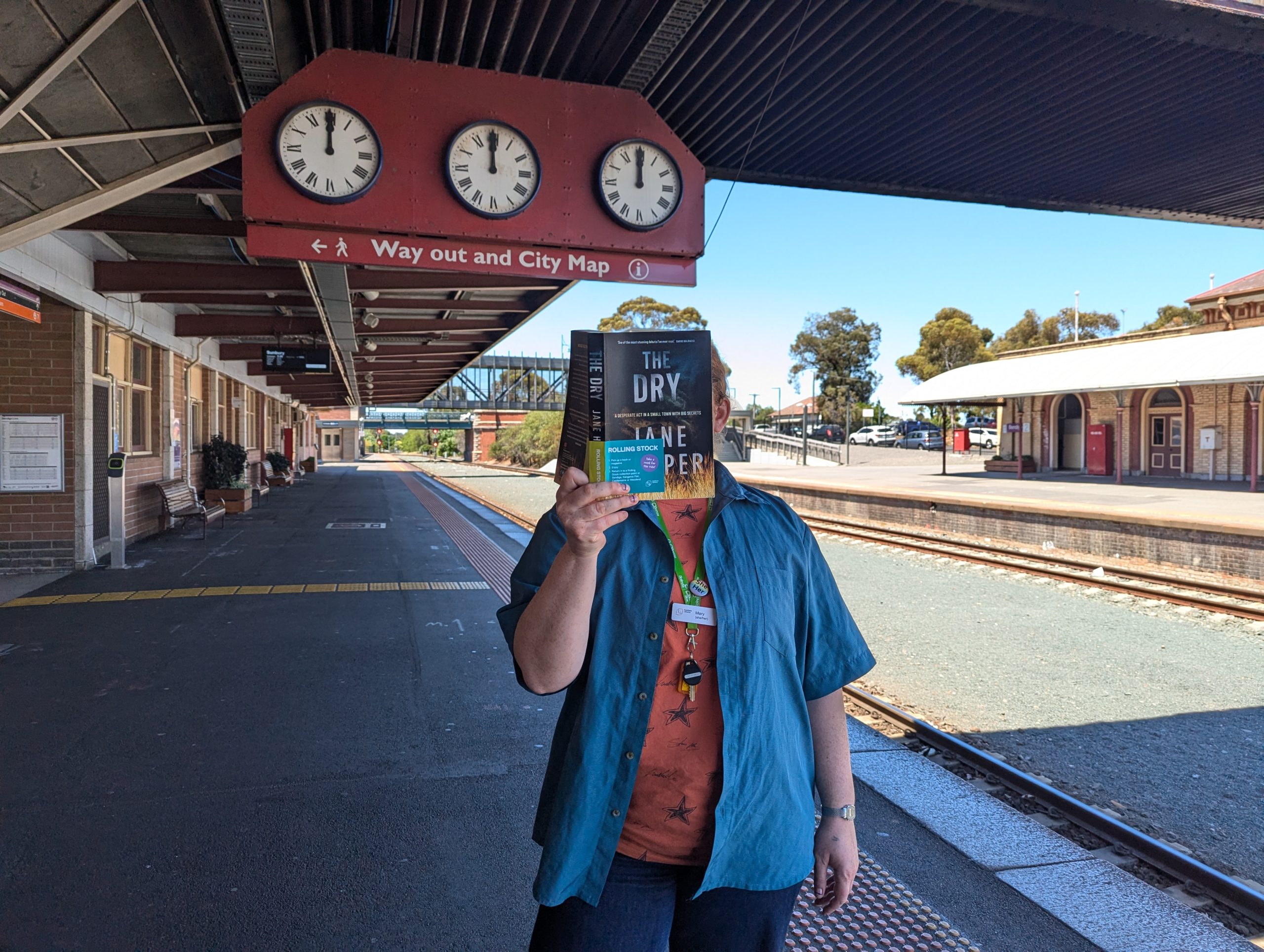 Woman holding Jane Harpers 'The Dry' in front of her face. She is standing on the Bendigo Train Station platform with the train lines in the background, and three clocks.